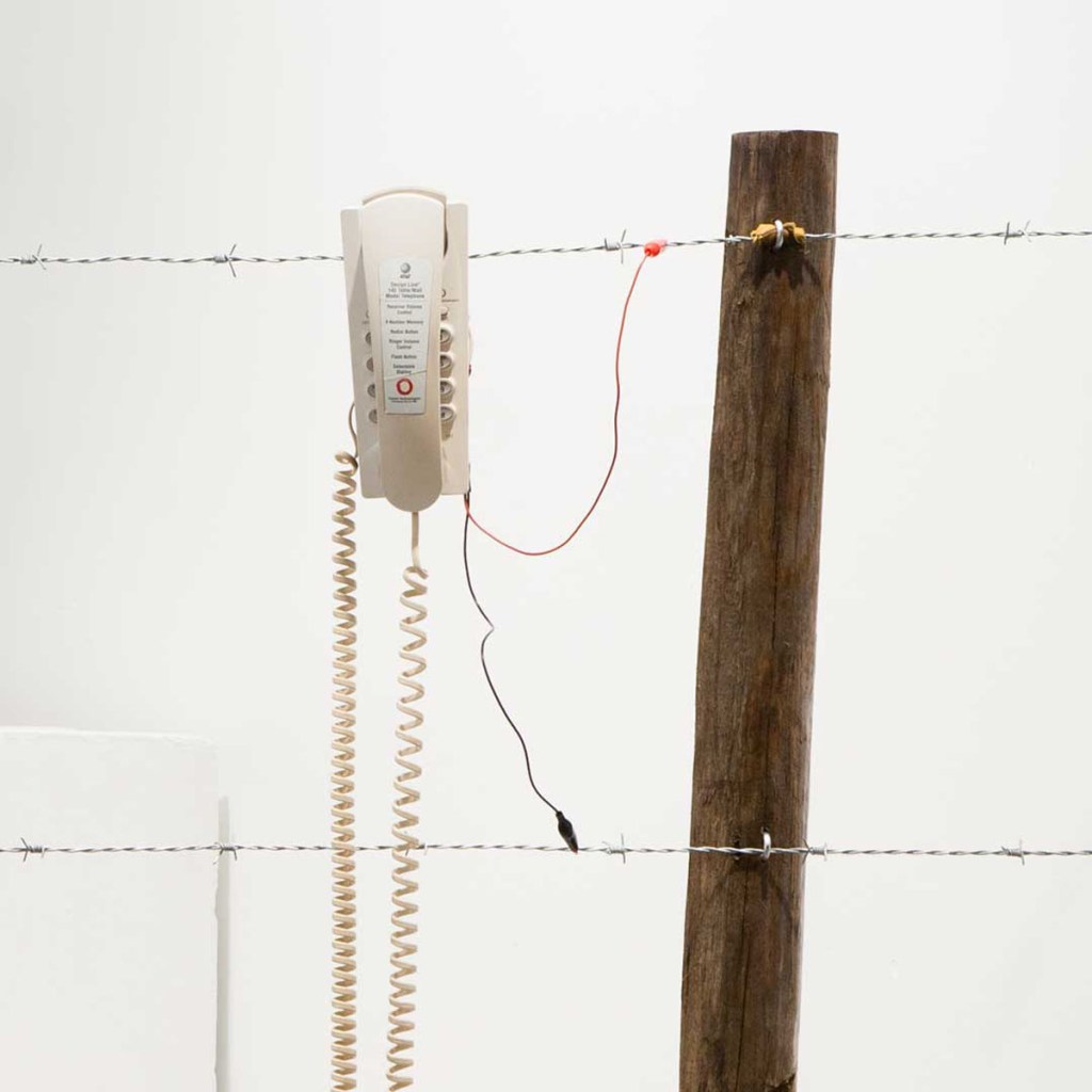 a fence post supports two strands of barbed wire. a telephone hangs from the top wire, and is connected by wires with alligator clips to both strands of wire. 